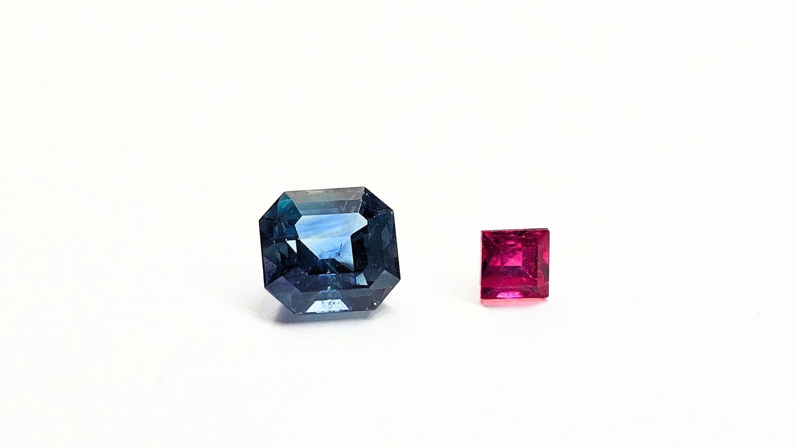 A natural blue sapphire and a red ruby, both step cut, side by side on a white background